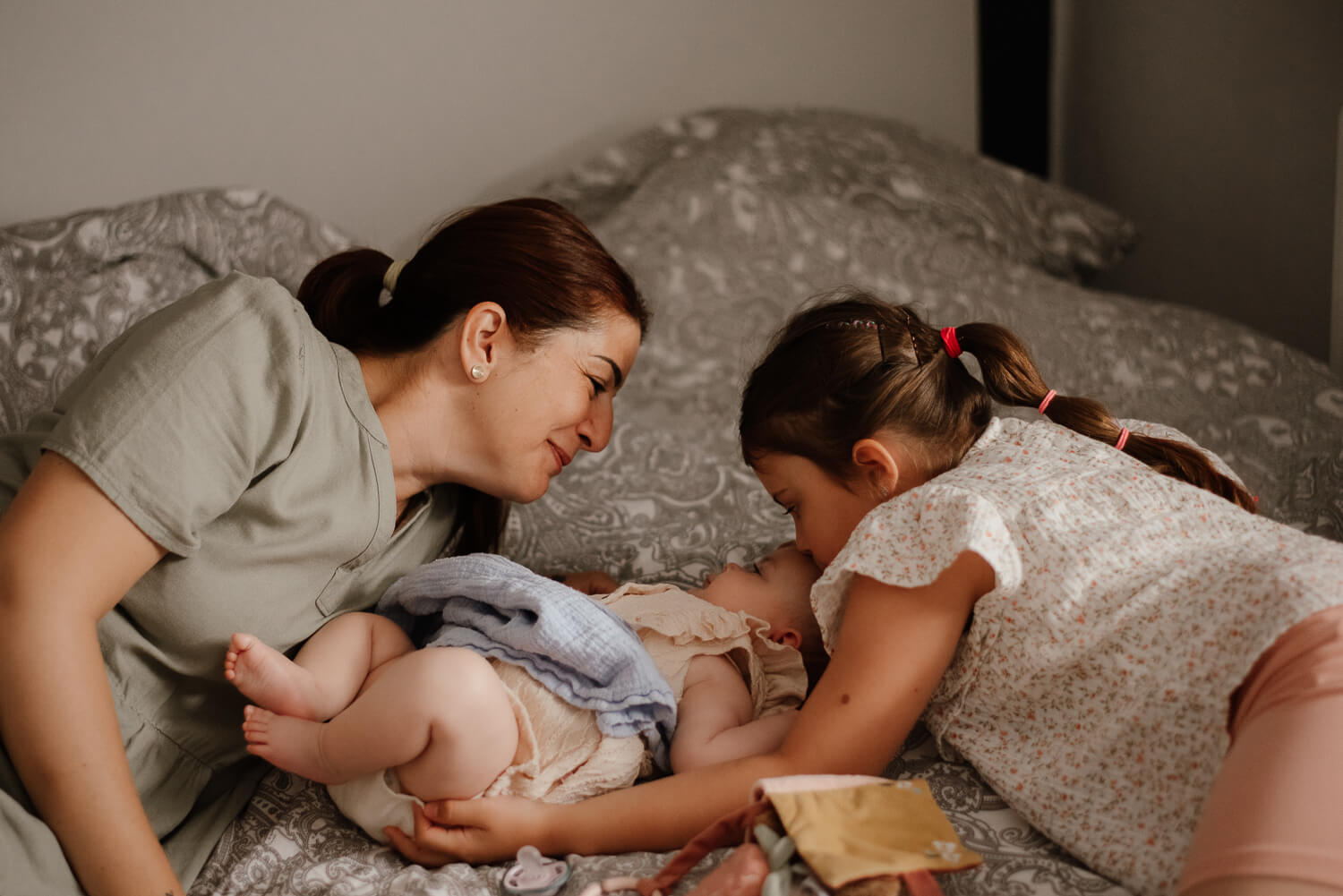 Mama mit Baby und Tochter auf dem Ehebett. Sie schaut glücklich zu ihrem Baby. Die große Schwester küsst die kleine Schwester.