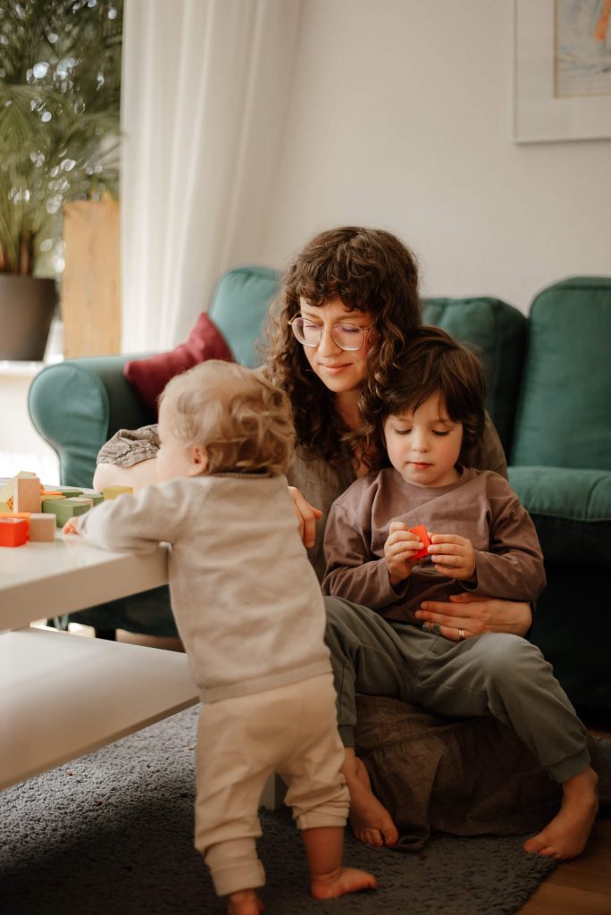 Mama sitzt auf dem Boden mit ihrem kleinen Sohn auf dem Schoss. Ein Baby hebt sich an den Couchtisch hoch.