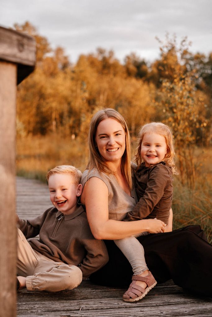 Mama mit ihren zwei Kindern, sitzend auf einem Holzweg. Alle 3 sehen sehr glücklich aus.
