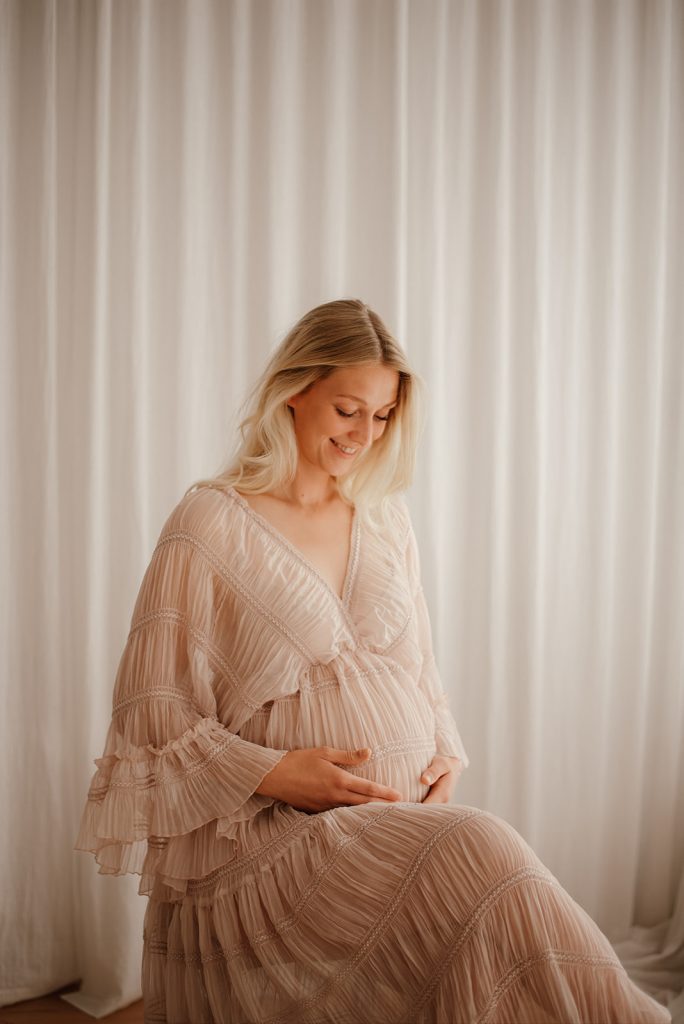 Schwangere Frau sitzt entspannt im Fotostudio und schaut zu ihrem Babybauch während eines natürlichen Schwangerschaftsshootings in Bordesholm bei Kiel.